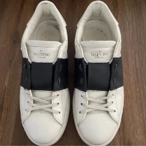 Valentino Garavani Open sneakers vit/svart byte!! - Snygga Valentino Garavani Open sneakers i vitt skinn med bred svart läderdetalj över mitten. Klassisk rund tå, vita snören och ikoniska nitar baktill. Perfekt för dig som gillar lyxiga och stilrena sneakers med en twist.