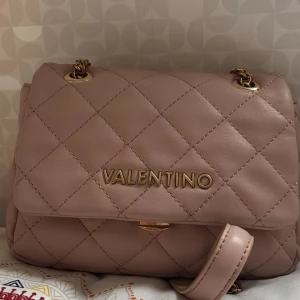 Quiltad axelväska från Valentino - Supersnygg quiltad axelväska från Valentino i ljusbeige skinn med guldfärgade kedjor och detaljer. Väskan har ett klassiskt rutmönster och en stilren design med Valentino-logga framtill. Perfekt storlek för det viktigaste och ger en lyxig touch till din outfit.