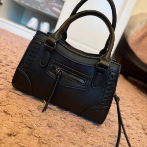 Svart axelväska från Gina Tricot - Snygg svart axelväska från Gina Tricot i skinnimitation med både handtag och justerbar axelrem. Väskan har dragkedja upptill, dekorativa sömmar och en liten ficka med dragkedja framtill. Perfekt storlek för mobilen, väskan finns ej att köpa längre på hemsidan .