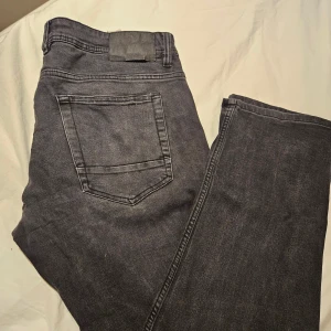 Svarta jeans från SMOG, 34/32 - Säljer ett par svarta jeans från SMOG i storlek 34/32. Klassisk femficksmodell med raka ben och normal passform. Jeansen är tillverkade i bomull och har en stilren look som funkar till det mesta. Perfekta för dig som gillar enkel och clean stil.