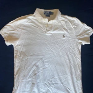 Vit pikétröja från Ralph Lauren - Klassisk vit pikétröja från Ralph Lauren med korta ärmar och broderad logga på bröstet. Tröjan har krage och knäppning med två knappar framtill. Tillverkad i bomull för en skön och luftig känsla. Perfekt för en clean och stilren look.