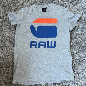 Grå G-Star RAW t-shirt med tryck - Snygg grå t-shirt från G-Star RAW med stort RAW-tryck i blått och orange på framsidan. Klassisk rund hals och korta ärmar. Perfekt för dig som gillar streetwear och vill ha en chill look. Mjuk bomull som känns skön mot huden.