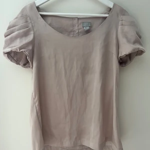 Ros blus med puffärm från H&M - Säljer en rosa blus från H&M i storlek 38. Blusen har puffiga korta ärmar med veckade detaljer och rund halsringning. Knappar i pärlemor på ryggen ger en extra touch. Materialet är mjukt och har en lätt glansig finish, perfekt för en chill och stilren look.