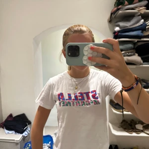 Vit Stella McCartney t-shirt med tryck - Säljer en vit t-shirt från Stella McCartney med färgglatt tryck på bröstet. Trycket har röda och blå leopardmönstrade bokstäver och gröna detaljer. Modellen är klassisk med rund hals och korta ärmar. Perfekt för dig som gillar designer och coola prints.