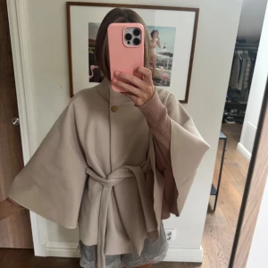 Beige poncho med bälte och vida ärmar - Snygg beige kappa/poncho med breda, vida ärmar och högt krage. Kappan har ett bälte i midjan som knyts och en dekorativ knapp vid halsen. Materialet ser ut att vara ull eller syntet, perfekt för dig som gillar stilrena och minimalistiska plagg.