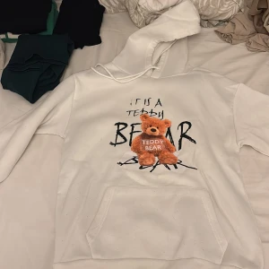 Vit hoodie med teddy bear-tryck - Vit hoodie med huva och känguruficka framtill. På bröstet finns ett stort tryck med texten 'IT IS A TEDDY BEAR' och en mjuk, brun nalle som 3D-detalj. Hoodien har dragsko i halsen och är långärmad. Perfekt för dig som gillar gulliga och unika plagg.
