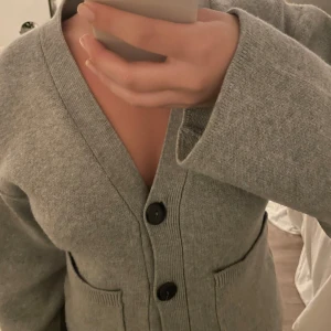 Grå kofta med stora knappar - Mysig grå kofta med v-ringning och tre stora svarta knappar framtill. Koftan har två fickor och långa ärmar med lite loose passform. Perfekt att slänga på sig över en t-shirt eller linne för en chill look.