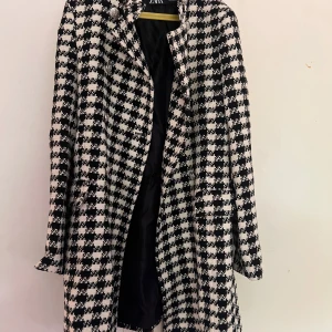 Rutig svartvit kappa från Zara XS - Säljer en snygg svartvit rutig kappa från Zara i storlek XS. Jackan har klassiskt hundtandsmönster, dekorativa silvriga knappar och är helfodrad. Perfekt för dig som vill sticka ut med en trendig och tidlös look.