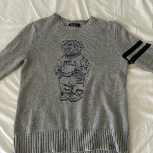 Grå stickad tröja Polo Bear Ralph Lauren - Grå stickad tröja från Polo Ralph Lauren med ikonisk Polo Bear broderad framtill. Tröjan har långa ärmar med två mörkblå ränder på ena ärmen och ribbade muddar. Perfekt för dig som gillar klassisk men ändå stilren stil.