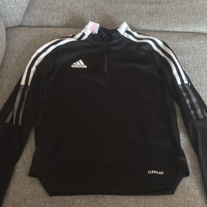 Svart Adidas half zip tröja - Svart långärmad Adidas-tröja med vita ränder längs ärmarna och logga på bröstet. Tröjan har half zip framtill och är gjord i ett lätt, syntetiskt material (AEROREADY) som andas. Perfekt för träning eller sportiga outfits.