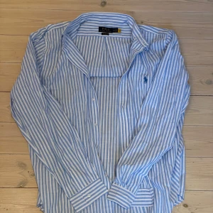 Blåvit randig skjorta Polo Ralph Lauren - Snygg blå och vit randig skjorta från Polo Ralph Lauren i klassisk passform. Skjortan har långa ärmar, knappar framtill och ikonisk broderad logga på bröstet. Tillverkad i tunn och skön bomull, perfekt för en stilren look.