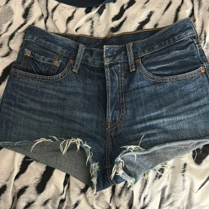 Levi's 501 blå jeansshorts med fransar - Snygga blå jeansshorts från Levi's modell 501 med klassisk femficksdesign och rå fransad kant. Shortsen har hög midja, slitna detaljer och tydliga kontrastsömmar. Perfekta för en chill och avslappnad stil under varma dagar.