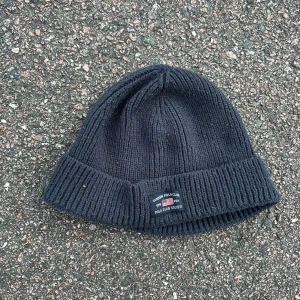 Mörkblå stickad mössa Vinson Polo Club - Snygg mörkblå stickad mössa från Vinson Polo Club med uppvikt kant och liten patch framtill. Perfekt för kyliga dagar och enkel att matcha med din jacka. Klassisk design som funkar till allt och ger en chill vibe.