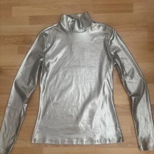  Ny.Silver polotröja från H&M - Säljer en glansig silverfärgad polotröja från H&M i storlek S. Tröjan har lång ärm och en hög krage som ger en cool och futuristisk vibe. Materialet är syntet och har en metallic finish som verkligen sticker ut. Perfekt för dig som vill ha något extra till outfiten.