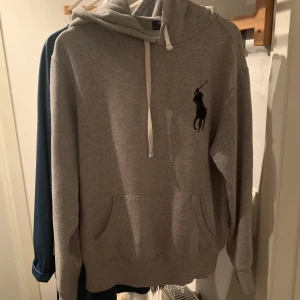 Grå hoodie från Polo Ralph Lauren - Snygg grå hoodie från Polo Ralph Lauren med klassisk svart logga på bröstet. Tröjan har huva med vit snörning och en stor magficka. Perfekt för dig som gillar en clean och stilren look. Mjuk bomullsblandning och normal passform.