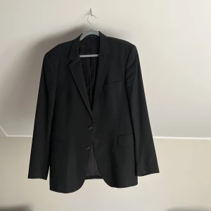 Svart kavaj från Tiger of Sweden - Snygg svart kavaj från Tiger of Sweden med klassisk passform och två knappar framtill. Kavajen har fickor med lock och en bröstficka, samt diskreta knappar vid ärmsluten. Perfekt för dig som vill ha en stilren och tidlös look.