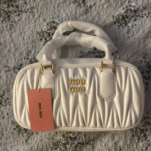 miu miu white bag - Supersnygg vit handväska från Miu Miu i quiltat skinn med guldiga detaljer och dragkedja. Väskan har ett stilrent och lyxigt utseende med dubbla handtag och logga framtill. Perfekt för dig som vill ha en trendig accessoar med exklusiv känsla.