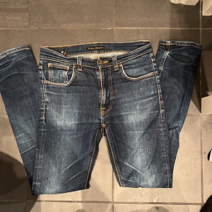 Mörkblå jeans från Nudie Jeans - Säljer ett par mörkblå Slim fit jeans från Nudie Jeans med klassisk femficksdesign och snygga slitningar. Jeansen har kontrastsömmar och den ikoniska Nudie-broderingen på bakfickan. Perfekta för dig som gillar en stilren och tidlös look.