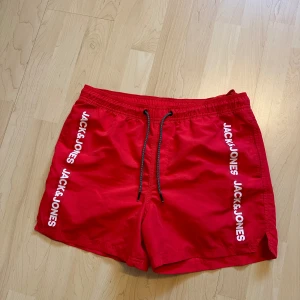 Röda badshorts Jack & Jones - Snygga röda badshorts från Jack & Jones med vit logga längs båda sidorna. Shortsen har elastisk midja med svart snörning och är perfekta för stranden eller poolen. Klassisk modell med sportig vibe.