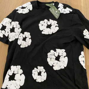 Svart t-shirt med blommor från Denim Tears - Svart t-shirt från Denim Tears med stora vita blommor tryckta över hela plagget. T-shirten har rund hals och korta ärmar, tillverkad i mjuk bomull. Snygg och unik design som sticker ut och passar perfekt till jeans eller shorts.