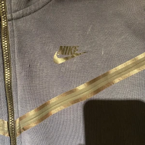 Nike zip hoodie med guldiga detaljer M - Nike hoodie i mörkgrått med dragkedja och guldiga ränder över bröstet. Klassisk Nike-logga i guld på vänster sida. Tillverkad i mjuk bomulls- och polyestermix. Perfekt för dig som gillar sportig och clean stil.