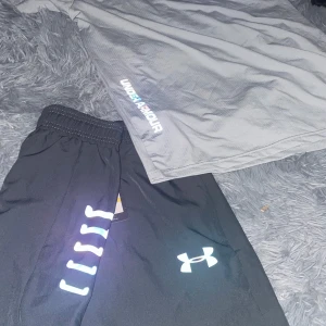 Under Armour - Snygga svarta träningsshorts från Under Armour med elastisk midja och reflekterande detaljer på sidan. Materialet är lätt och snabbtorkande polyester, perfekt för intensiva pass. Loggan syns tydligt framtill och shortsen har en loose passform.