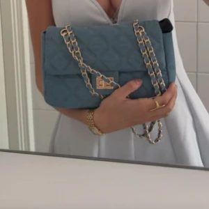 Blå quiltad axelväska med guldkedja - Säljer en snygg blå quiltad axelväska med guldfärgad kedja och vridlås framtill. Väskan har en stilren design och är perfekt för att lyfta vilken outfit som helst. Kedjan kan bäras både enkel och dubbel för olika look.