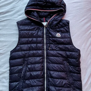 Moncler väst - Snygg svart dunväst från Moncler med quiltad design och huva. Västen har dragkedja framtill, två fickor med dragkedja och Moncler-logga på bröstet. Perfekt för lager-på-lager och streetstyle. Materialet är glansigt och lätt, fylld med äkta dun.