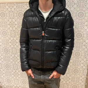 Svart pufferjacka från Save The Duck - Säljer en svart pufferjacka från Save The Duck med huva och dragkedja framtill. Jackan är vadderad och har en glansig finish, samt Save The Duck-logga på ärmen. Tillverkad i nylon och polyester, perfekt för kyliga dagar.