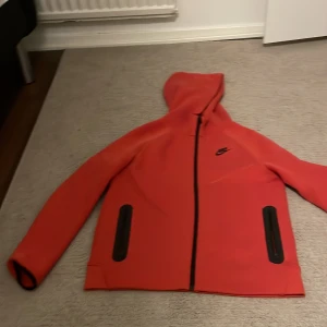 Röd Nike vindjacka med huva XL - Nike vindjacka i rött med svart dragkedja och svarta detaljer vid fickorna. Jackan har huva, hel dragkedja och sportig look. Perfekt för dig som gillar streetstyle och vill ha något bekvämt och snyggt. Passar dig som vill sticka ut med färg.