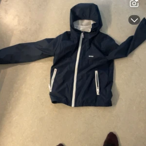 Mörkblå vindjacka från BOSS med huva (barnstorlek) - Snygg mörkblå vindjacka från BOSS med vit dragkedja och två fickor framtill. Jackan har huva och meshfoder på insidan för extra komfort. Perfekt för blåsiga dagar och enkel att matcha med olika outfits. Ifall du har någon lillebror eller liknande 