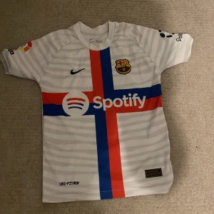 Barcelona Ansu Fati 10 vit matchtröja - Säljer en Barcelona fotbollströja med Ansu Fati och nummer 10 på ryggen. Tröjan är vit med ljusgråa horisontella ränder, har ett stort kors i rött och blått på framsidan, samt Spotify och Polkadot som sponsorer. Märkt med Nike Dri-FIT ADV och klubbmärke på bröstet.