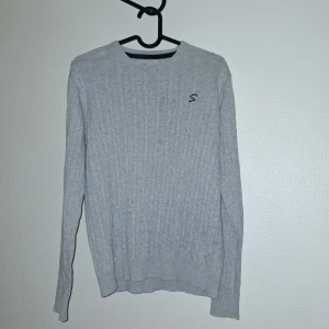 Ljusgrå stickad tröja från Skill - Snygg ljusgrå stickad tröja från Skill med diskret broderad logga på bröstet. Tröjan har ribbade muddar och ett klassiskt rakt snitt. Perfekt för dig som gillar enkel och stilren look.