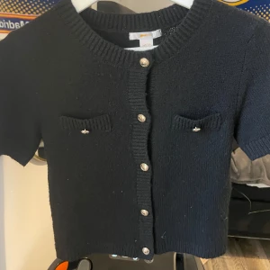 Svart stickad kofta  - Kort svart stickad kofta från Gina Tricot med rund halsringning, korta ärmar och dekorativa knappar framtill. Två små fickor med snygga detaljer på bröstet. Perfekt att slänga över en topp för en chill look.