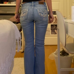 Ljusblå bootcut jeans med broderi - Säljer ett par ljusblå bootcut jeans från Y Jeans med snygga broderade bakfickor. Fickorna har detaljer med kors, vingar och glittriga pärlor. Jeansen har normal passform och är tillverkade i klassiskt denimtyg. Perfekta för dig som vill sticka ut lite extra!