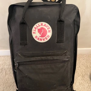 Svart Fjällräven Kånken Laptop 15” ryggsäck - Klassisk svart Fjällräven Kånken ryggsäck med ikonisk rund logga framtill. Rymligt huvudfack, framficka med dragkedja och justerbara axelremmar. Tillverkad i slitstarkt material, perfekt för skolan eller cityhäng. Sittunderlag ingår, väskan är hel.