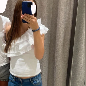 Vit oneshoulder volangtopp. Mycket efterfrågad - Säljer en vit oneshoulder topp med stora volanger över axeln. Toppen är figurnära och har en snygg asymmetrisk design som ger en trendig vibe. Perfekt att styla med jeans eller kjol för en cool look. Använd max tre gånger, har ingen användning av den. Men annars väldigt fin.