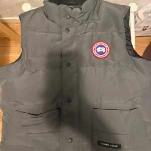 Grå dunväst från Canada Goose M - Säljer en grå dunväst från Canada Goose i storlek M. Västen har hög krage, två stora fickor framtill och klassisk logga på bröstet. Stängs med tryckknappar och har en stilren, enkel design. Perfekt för lager-på-lager under kyliga dagar.