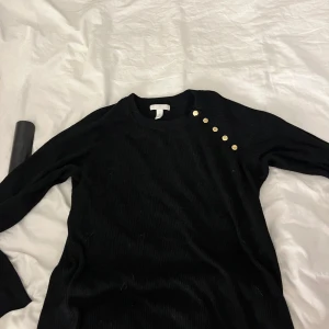 Svart ribbad tröja med guldknappar H&M - Svart ribbad tröja från H&M i storlek L men passar nu S, XS. Tröjan har lång ärm och snygga guldknappar på ena axeln som ger en extra touch. Perfekt för dig som gillar stilrena plagg med en twist.