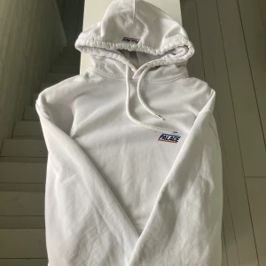 Vit hoodie från Palace med logga - Säljer en clean vit hoodie från Palace med broderad logga i blått och rött på bröstet. Tröjan har huva med snörning och är tillverkad i mjuk bomull. Perfekt för dig som gillar streetwear och vill ha en stilren look.
