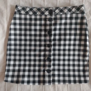 Svartvit rutig kort kjol med knappar - Supersnygg svartvit rutig kjol med knappar framtill. Kjolen är i mjukt stretchigt bomullstyg. Storlek 40. I fint skick.