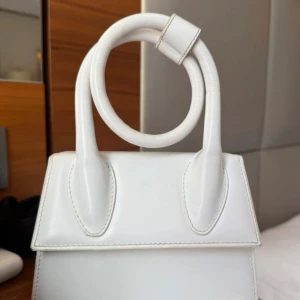 Vit Jacquemus väska - Säljer en stilren vit handväska med unikt rundat handtag och guldfärgade metalldetaljer. Väskan har en minimalistisk design med slät yta och är tillverkad i skinn. Perfekt för dig som gillar trendiga accessoarer med en twist.