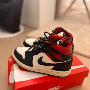 Nike Air Jordan 1 svart/röd/vit 38 - Nike Air Jordan 1 sneakers i klassisk svart, röd och vit färgkombination. Använda engång ser ut helt nya. Skorna har högt skaft, svart swoosh, röda detaljer och ikonisk Air Jordan-logga på sidan. Tillverkade i skinn och textil med snörning och rund tå. Perfekta för dig som vill sticka ut med en tidlös streetstil. Boxen finns ej med men genom QR koden och serial nummer i skorna kan du identifiera att de är äkta