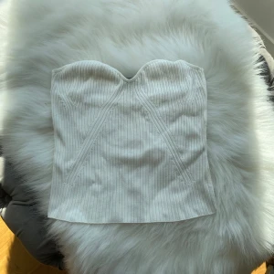 Vit ribbad korsettopp från Lindex - Säljer en snygg vit korsettopp från Lindex i ribbad bomull. Toppen är axelbandslös med markerade sömmar framtill som ger en cool form. Perfekt att styla med jeans eller kjol för en trendig look.