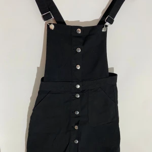 Svart kort jeans klänning med hängslen - Säljer en svart kort jeans klänning med hängslen och knappar framtill. Klänningen har två fickor och justerbara axelband i klassisk dungare-stil. Perfekt för dig som gillar en enkel och cool look. 