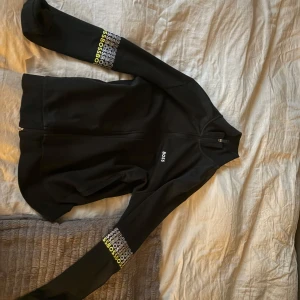 Svart ziptröja från BOSS  - Svart långärmad ziptröja från BOSS med vit logga på bröstet och coolt BOSS-tryck i vitt och gult på båda ärmarna. Tröjan har hög krage och hel dragkedja framtill. Perfekt för dig som gillar stilren streetwear med en twist.