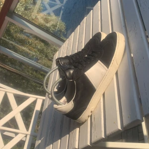 Valentino Garavani svarta sneakers - Snygga svarta och vita sneakers från Valentino Garavani med klassisk design. Skorna har svarta mockadetaljer, vita skinnpartier och coola nitar baktill på sulan. Perfekta för dig som vill ha en stilren och exklusiv look.