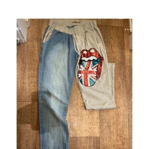 Unika tvåfärgade byxor med tryck - Säljer ett par riktigt coola byxor med ena benet i ljusblå denim och andra i grå bomull. På det grå benet finns ett stort tryck med läppar och Union Jack-flagga. Byxorna har resår i midjan och avslappnad passform. Perfekta för dig som vill sticka ut!