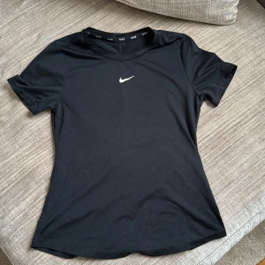 Svart Nike t-shirt i sportmaterial - Svart t-shirt från Nike med klassisk vit swoosh-logga på bröstet. Gjord i ett mjukt och stretchigt material som andas, perfekt för träning. Kortärmad och figurnära passform med rund halsringning. Snygg och enkel design med diskreta sömmar.