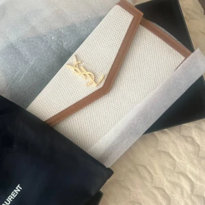 Saint Laurent kuvertväska i beige canvas - Snygg kuvertväska från Saint Laurent i beige canvas med bruna skinnkanter och guldigt YSL-emblem framtill. Stilren och lyxig design som passar perfekt till en trendig look. Väskan har en klassisk kuvertform och exklusiva detaljer. Använt fåtal gånger. Små repor på loggan de syns knappt. Därför har jag sänkt priset för fler bilder skriv